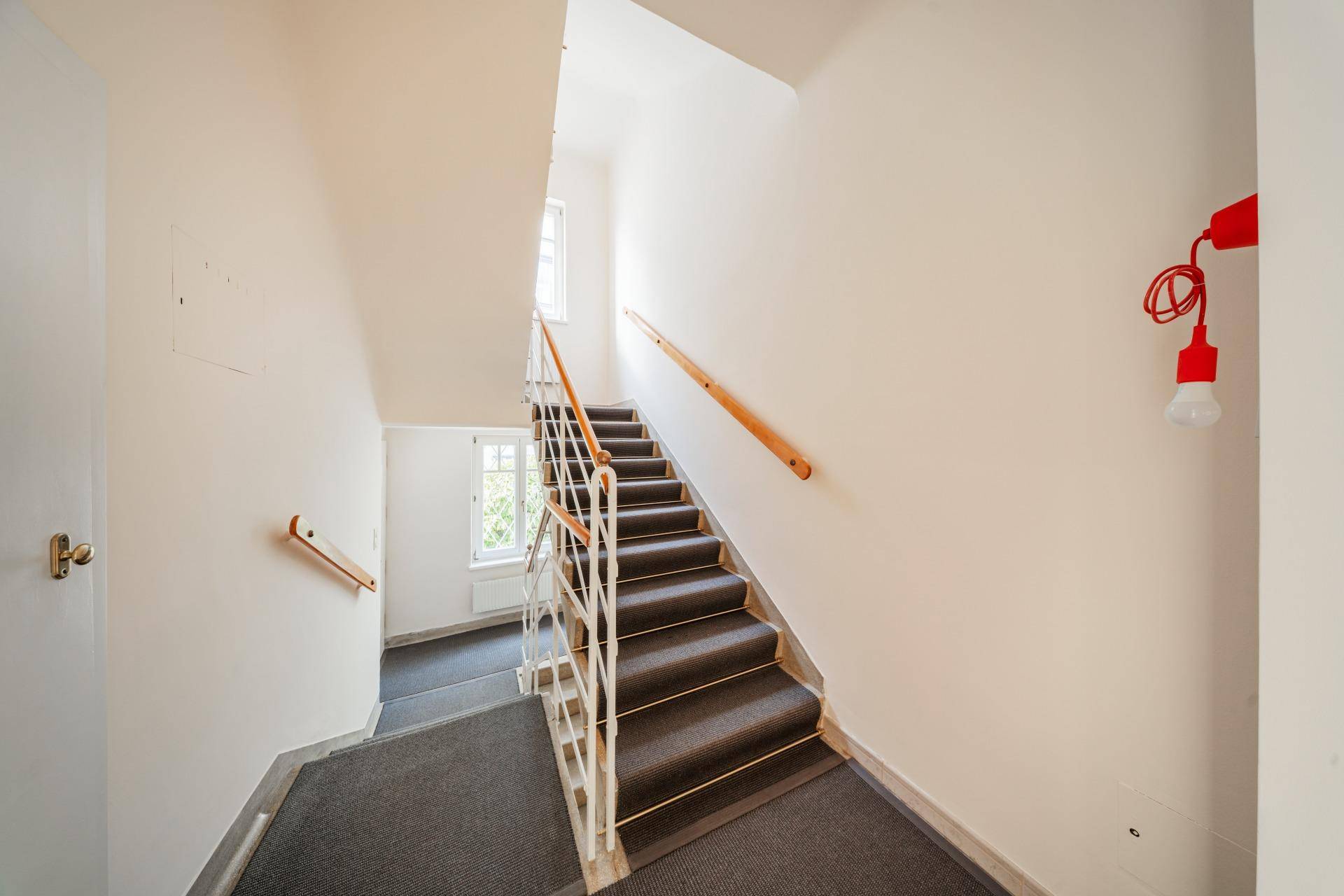 Helles Treppenhaus mit Teppichboden, Holzgeländer und Fenstern für natürliches Licht in den Etagen.