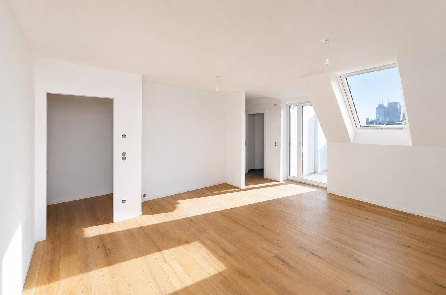 Heller, leerer Raum im Dachgeschoss mit Holzboden, großem Dachfenster und Zugang zum Balkon mit Stadtblick.