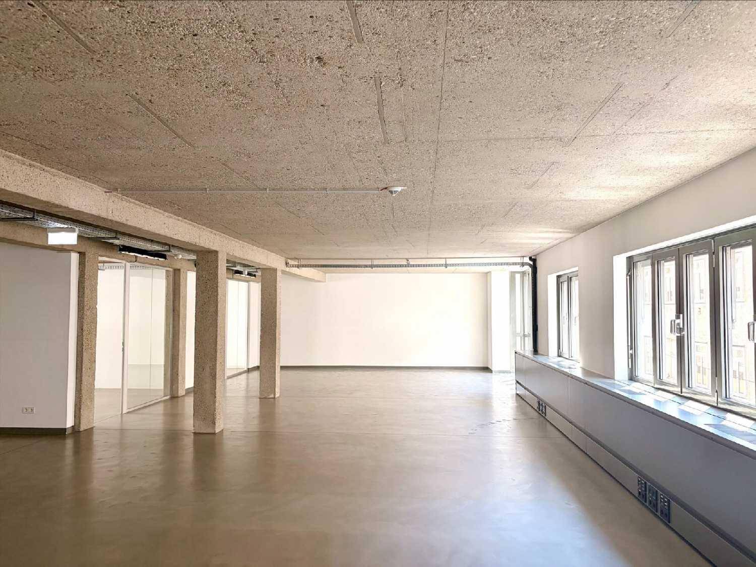 Geräumiger, leerer Bürobereich mit großen Fenstern, Betonboden und unverputzter Decke.