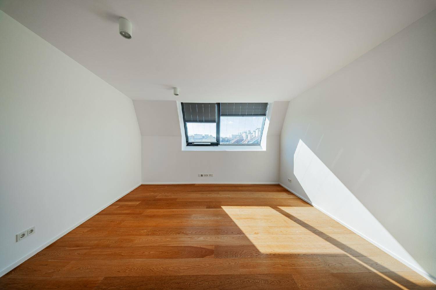 Leeres Schlafzimmer mit Holzboden, weißen Wänden und einem Fenster, das viel Licht hereinlässt.