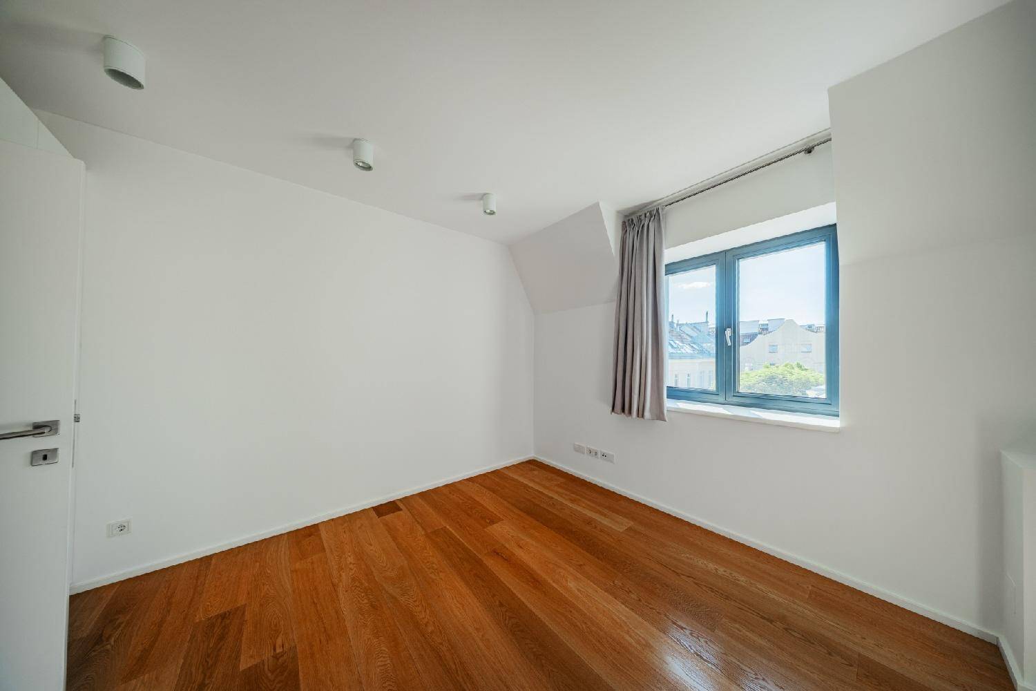 Helles Schlafzimmer mit Holzboden, weißen Wänden und einem Fenster mit Blick auf die Umgebung.