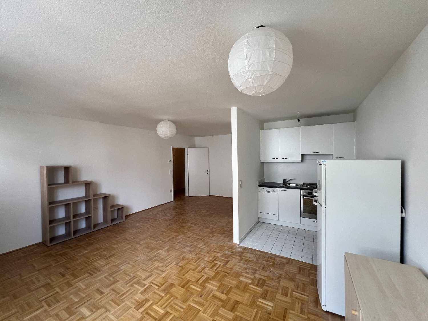 Offener Wohnbereich mit Parkettboden und integrierter Küchenzeile mit weißen Schränken und Kühlschrank.