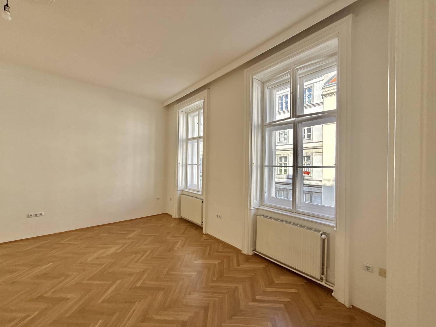 Geräumiges Zimmer mit Fischgrätparkett und hohen Fenstern, die viel Tageslicht hereinlassen und einen Blick nach draußen bieten.