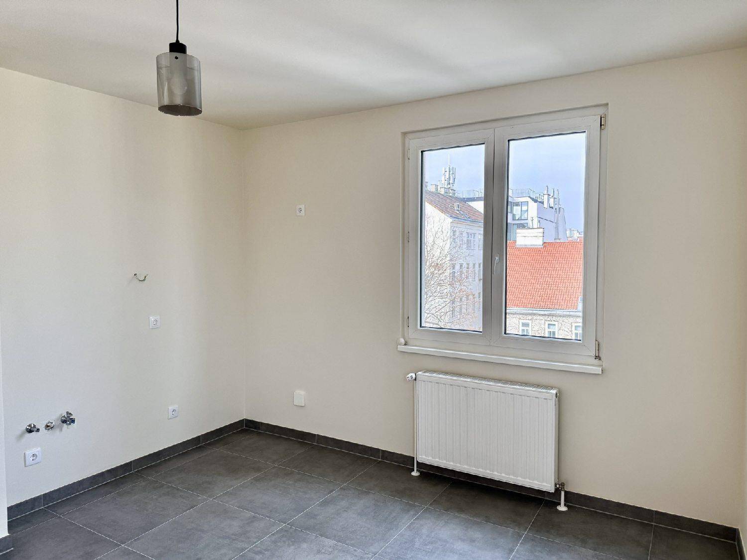 Leerer Raum mit grauen Fliesen, Fenster, Heizkörper und sichtbaren Wasseranschlüssen.