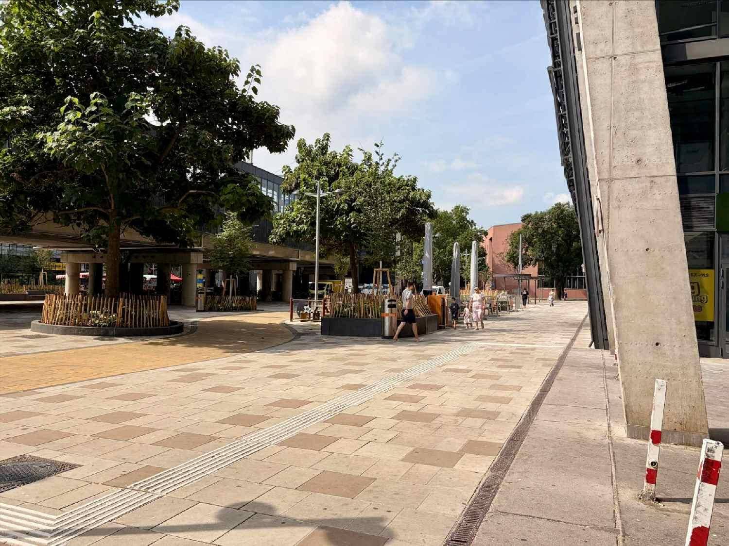 Weitläufiger öffentlicher Platz mit Bäumen, Sitzgelegenheiten und Passanten vor dem modernen Gebäude.