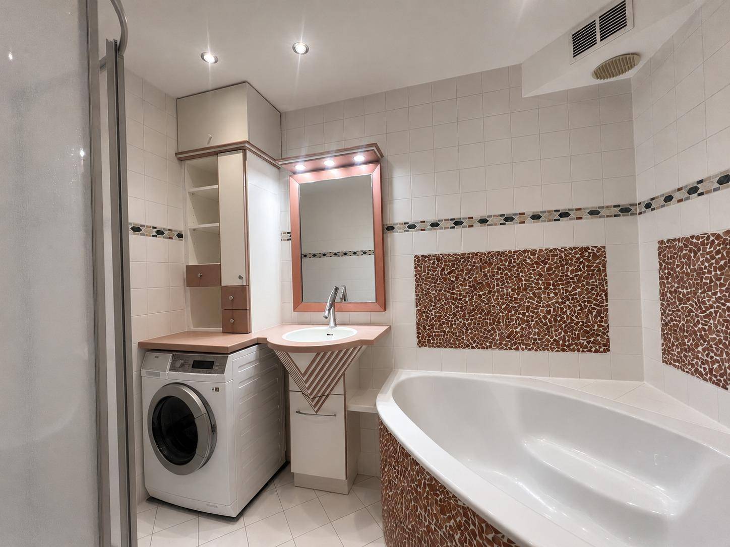 Badezimmer mit Badewanne, Waschbecken, Spiegel und einer Waschmaschine, ausgestattet mit gemusterten Fliesen.