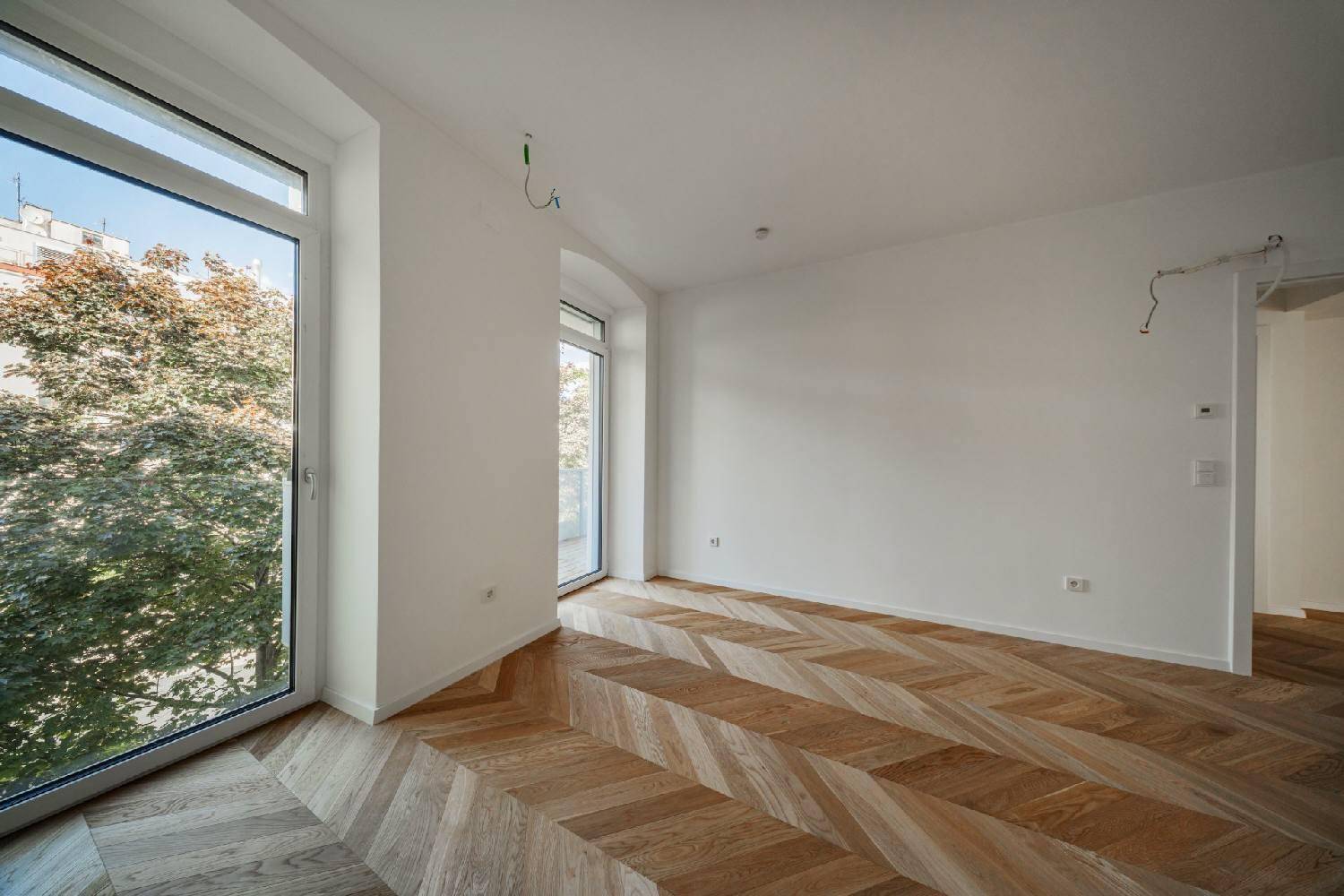 Eckbereich eines hellen Zimmers mit großem Fenster und Blick ins Grüne, ausgestattet mit Fischgrätparkett.