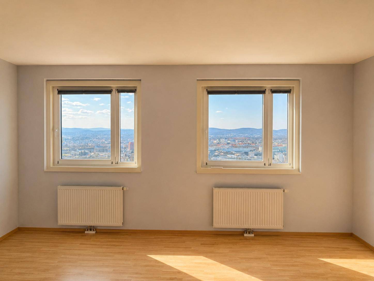 Zwei große Fenster in einem hellen Raum mit Parkettboden und Heizkörpern unter den Fenstern.