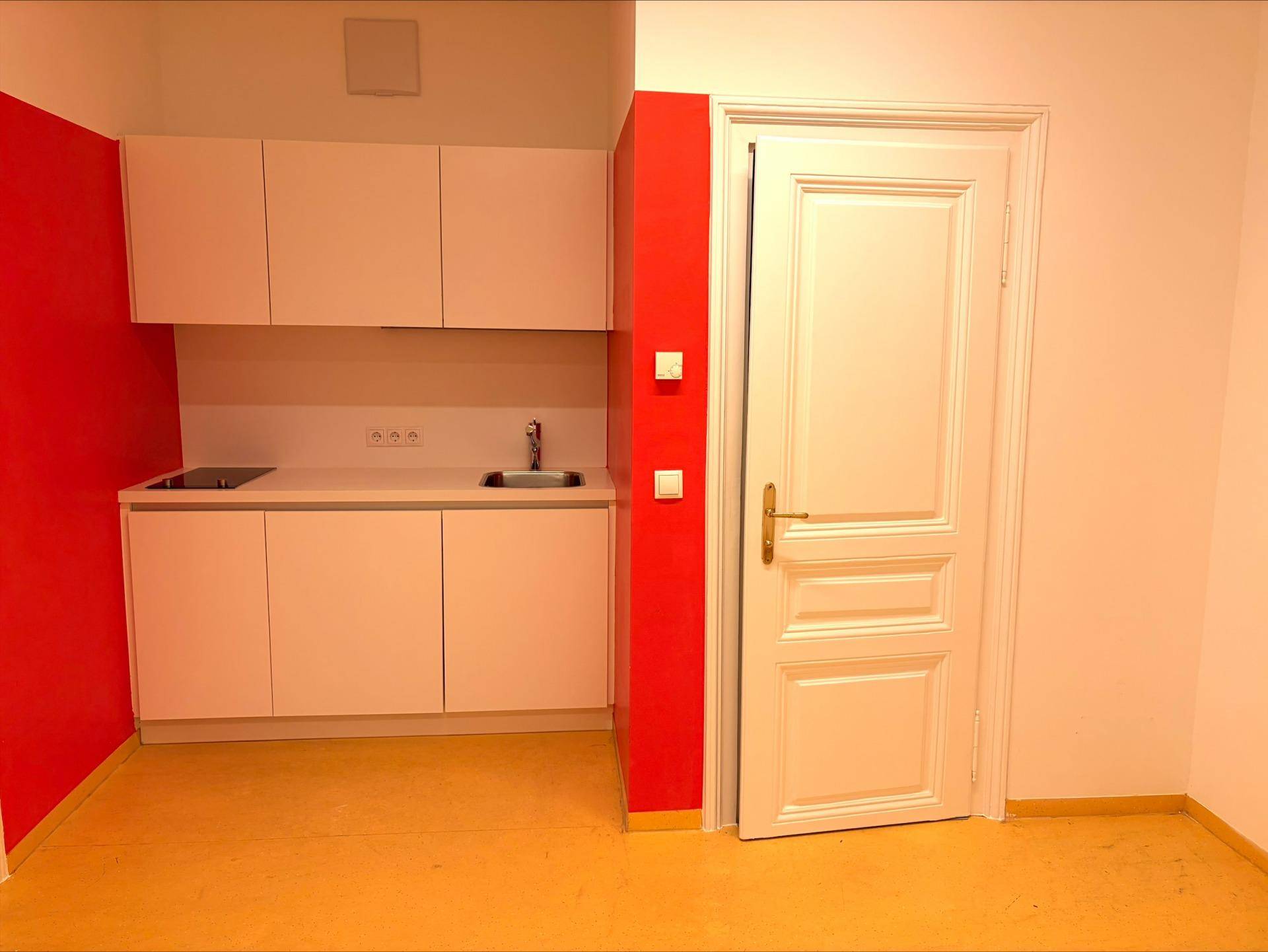 Kleine, moderne Küchenzeile mit weißen Schränken, Spüle und Kochfeld vor einer roten Wand.