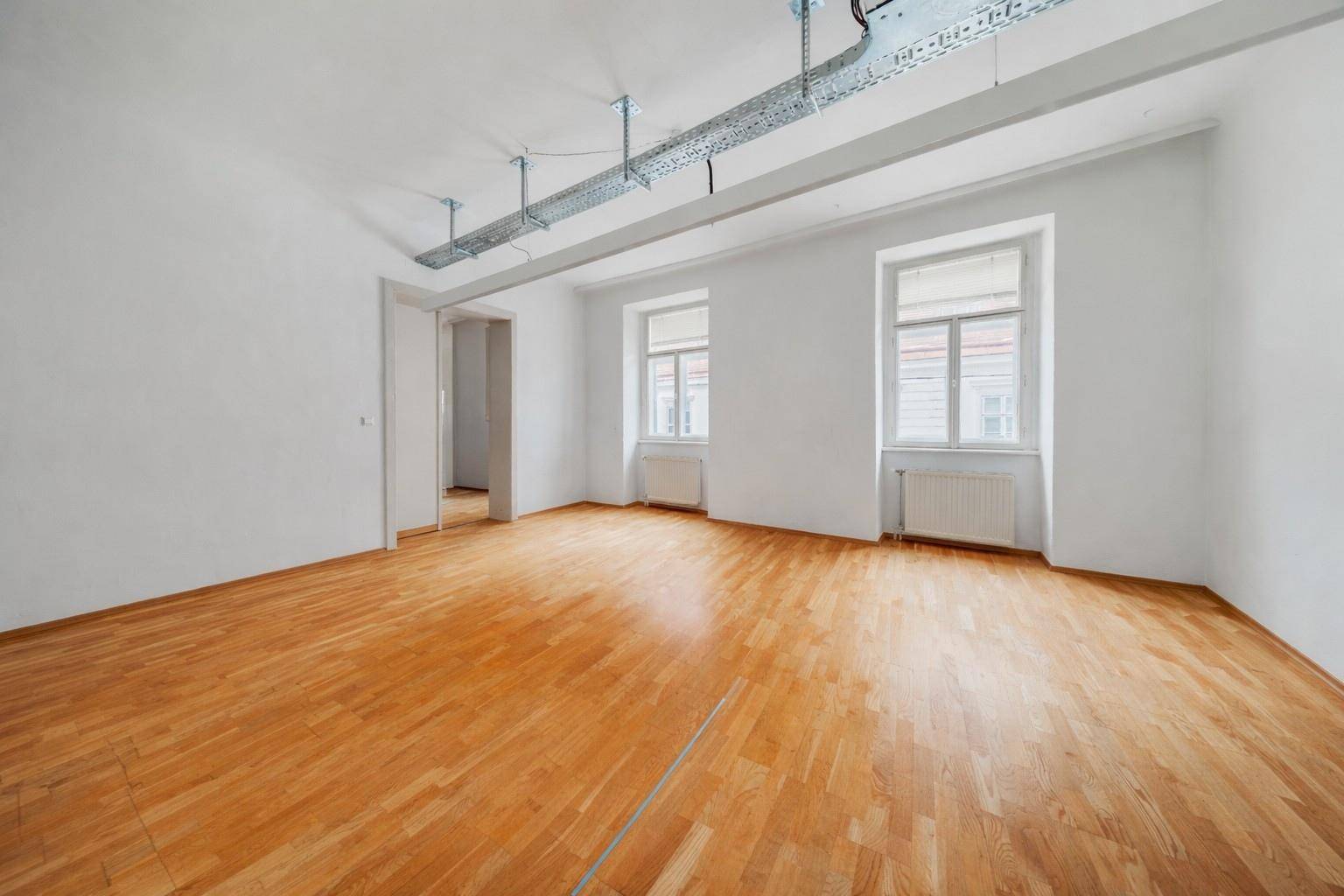 Heller Büroraum mit zwei großen Fenstern, Holzboden und sichtbaren Kabelkanälen an der Decke, frisch renoviert.