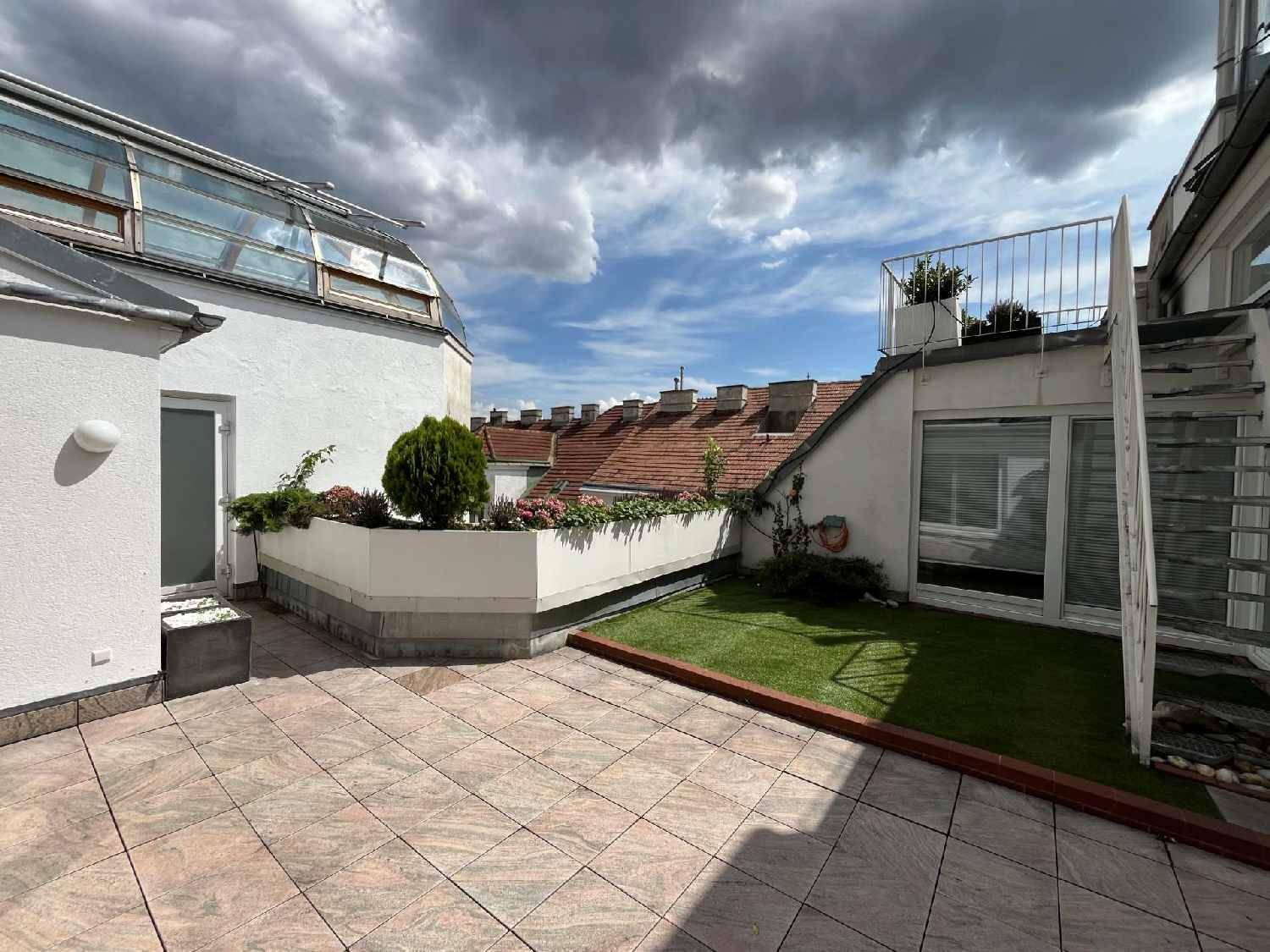 Weitläufige Dachterrasse mit Fliesenboden, Grünflächen und Blick über die Dächer der Stadt.