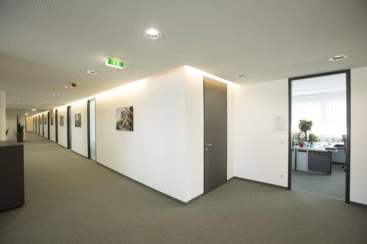Langer Bürogang mit Teppichboden, weißen Wänden und Glastüren, die zu einzelnen Büros führen.