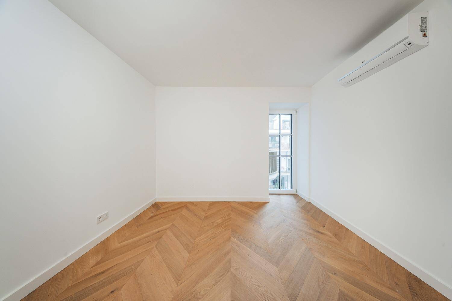 Kleinerer Raum mit Fischgrätparkett, Klimaanlage und einem hohen Fenster.