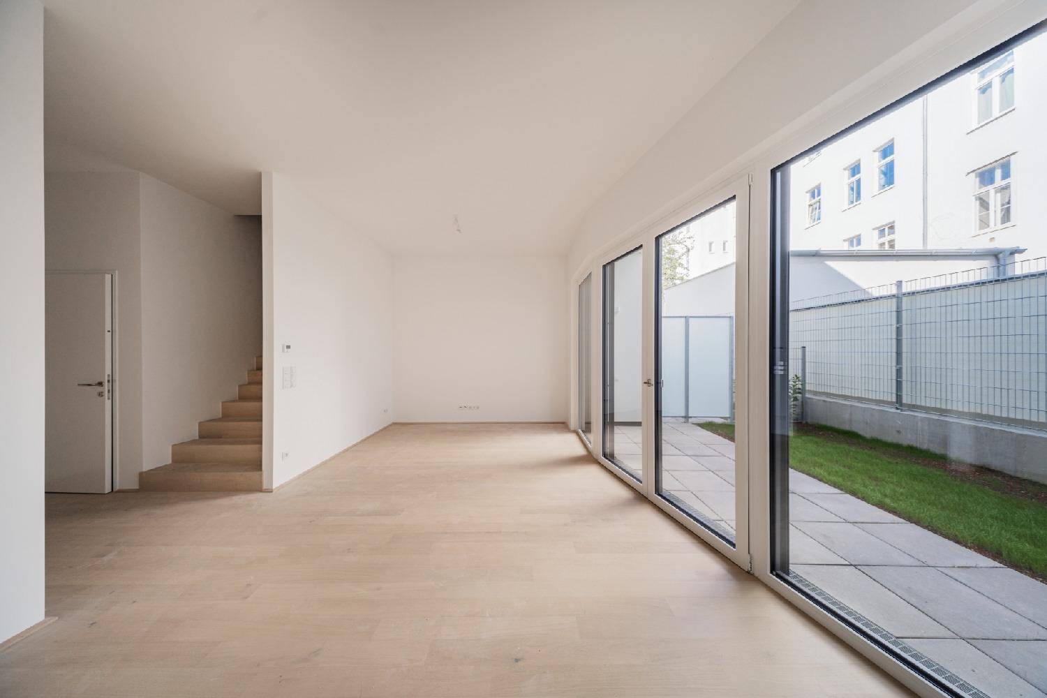 Heller, leerer Wohnbereich mit großen Fenstern und Zugang zur Terrasse und zum Garten.