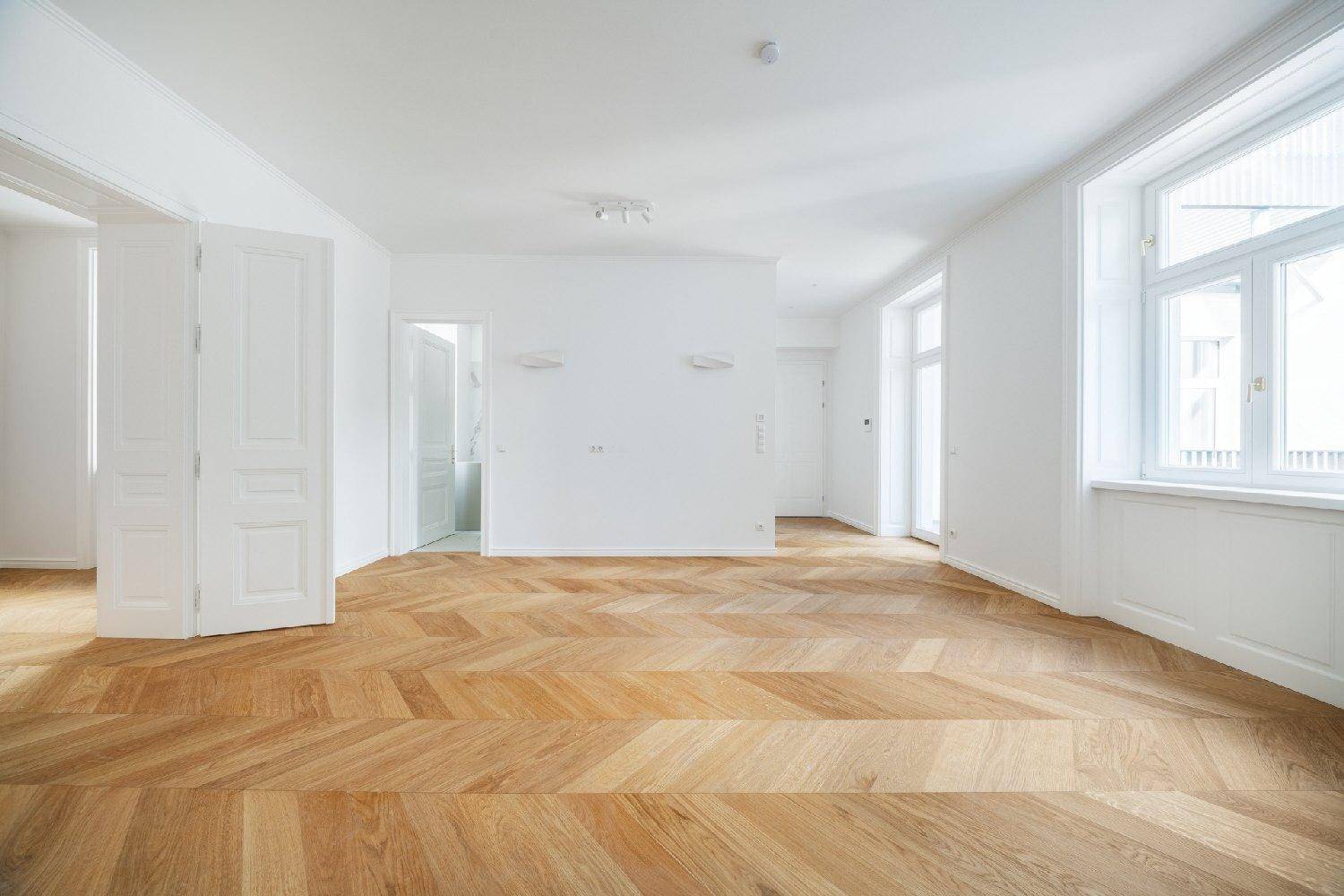 Geräumiges, helles Zimmer mit elegantem Fischgrätparkett und weißen Wänden, ideal für Wohnzwecke.
