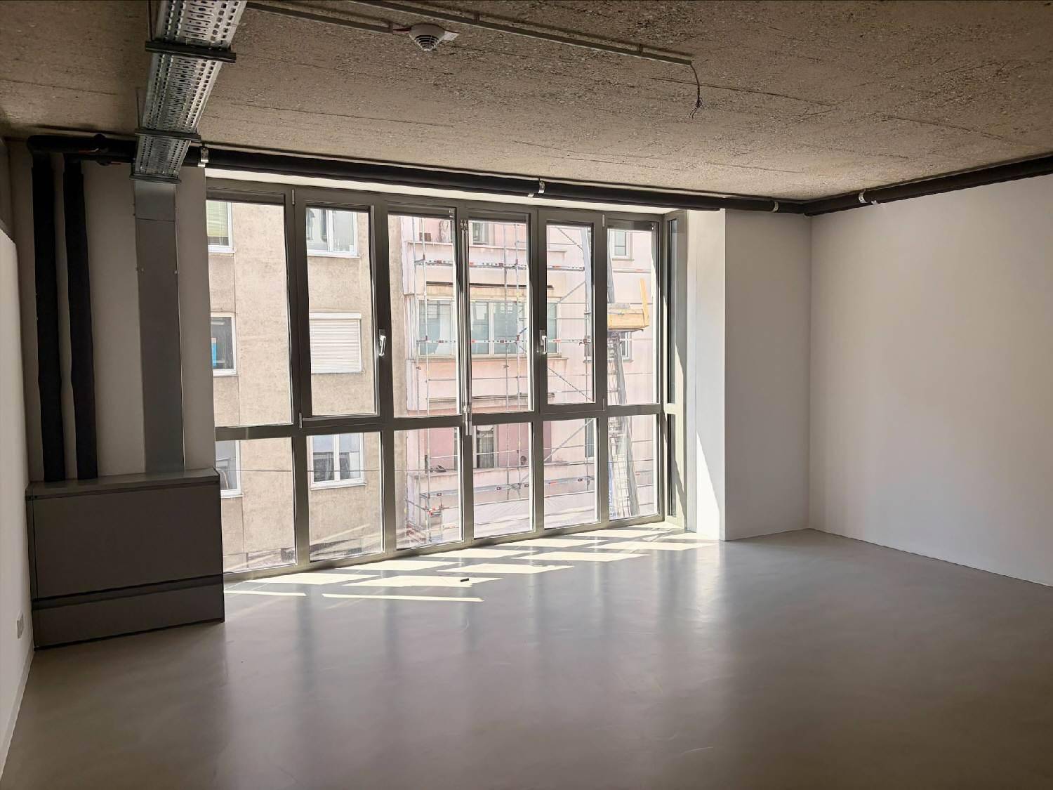 Heller Büroraum mit großer Fensterfront, Betonboden und Blick auf die Stadt.