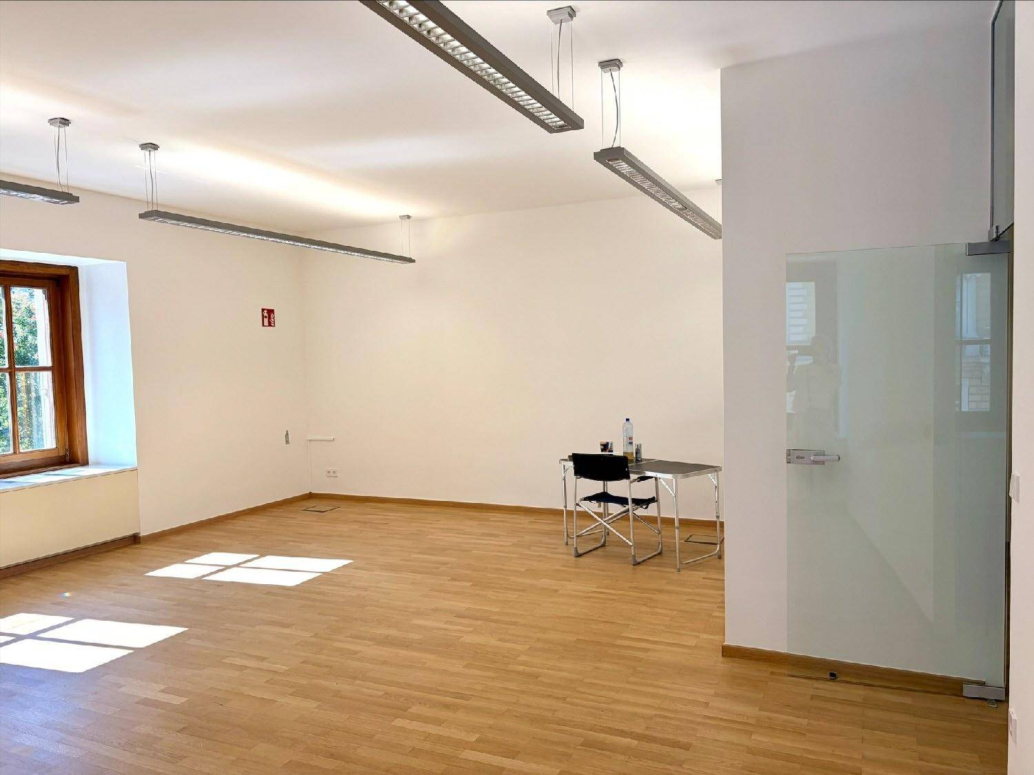 Heller Raum mit Holzboden, weißen Wänden und einer modernen Glastür, teilweise möbliert mit einem kleinen Tisch.