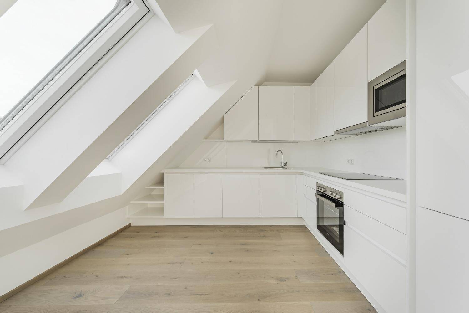 Moderne weiße Küche mit Dachschrägen, integrierten Geräten und hellem Holzboden.