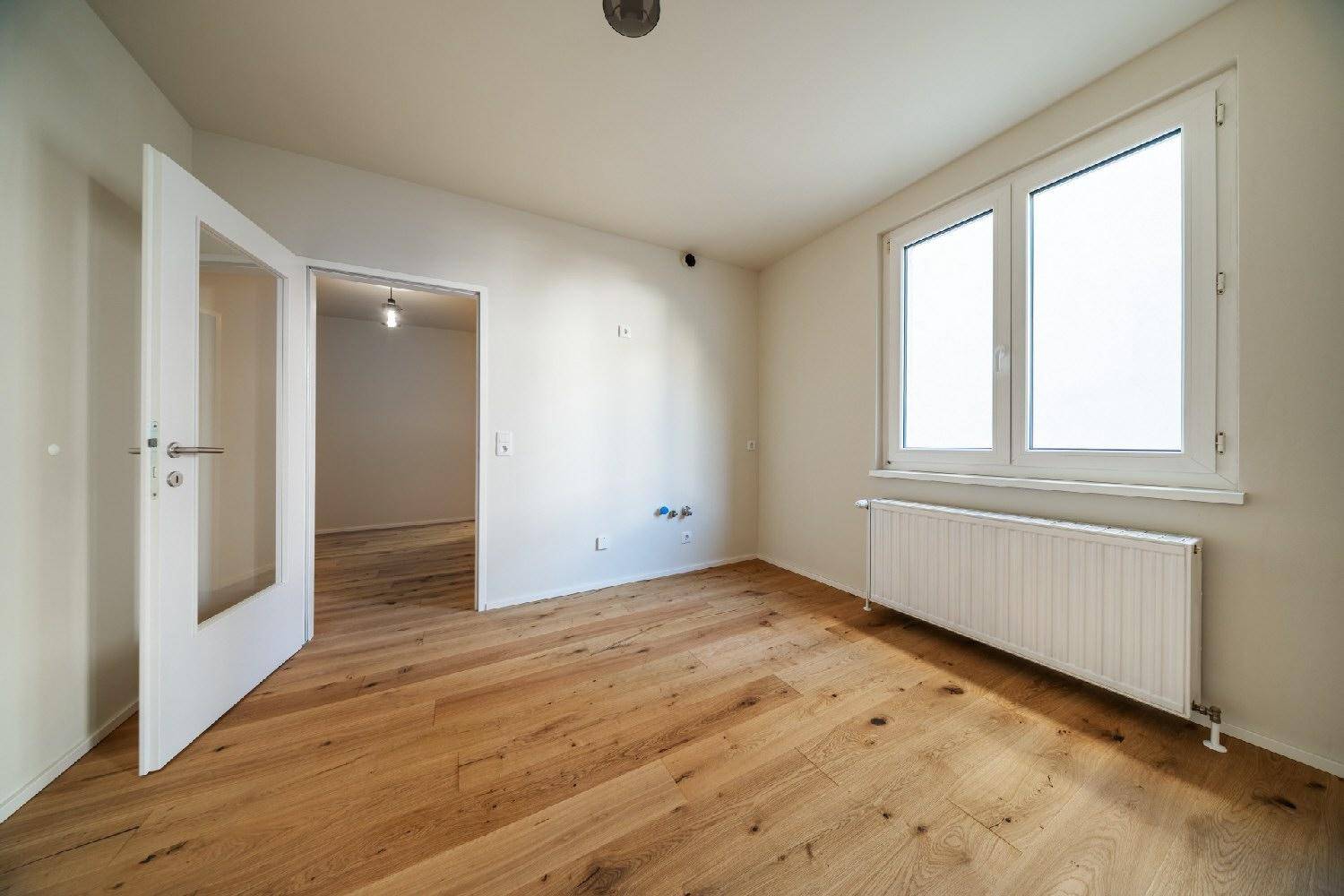 Helles Zimmer mit Holzboden, einem Fenster und einer offenen Tür zu einem weiteren Raum.