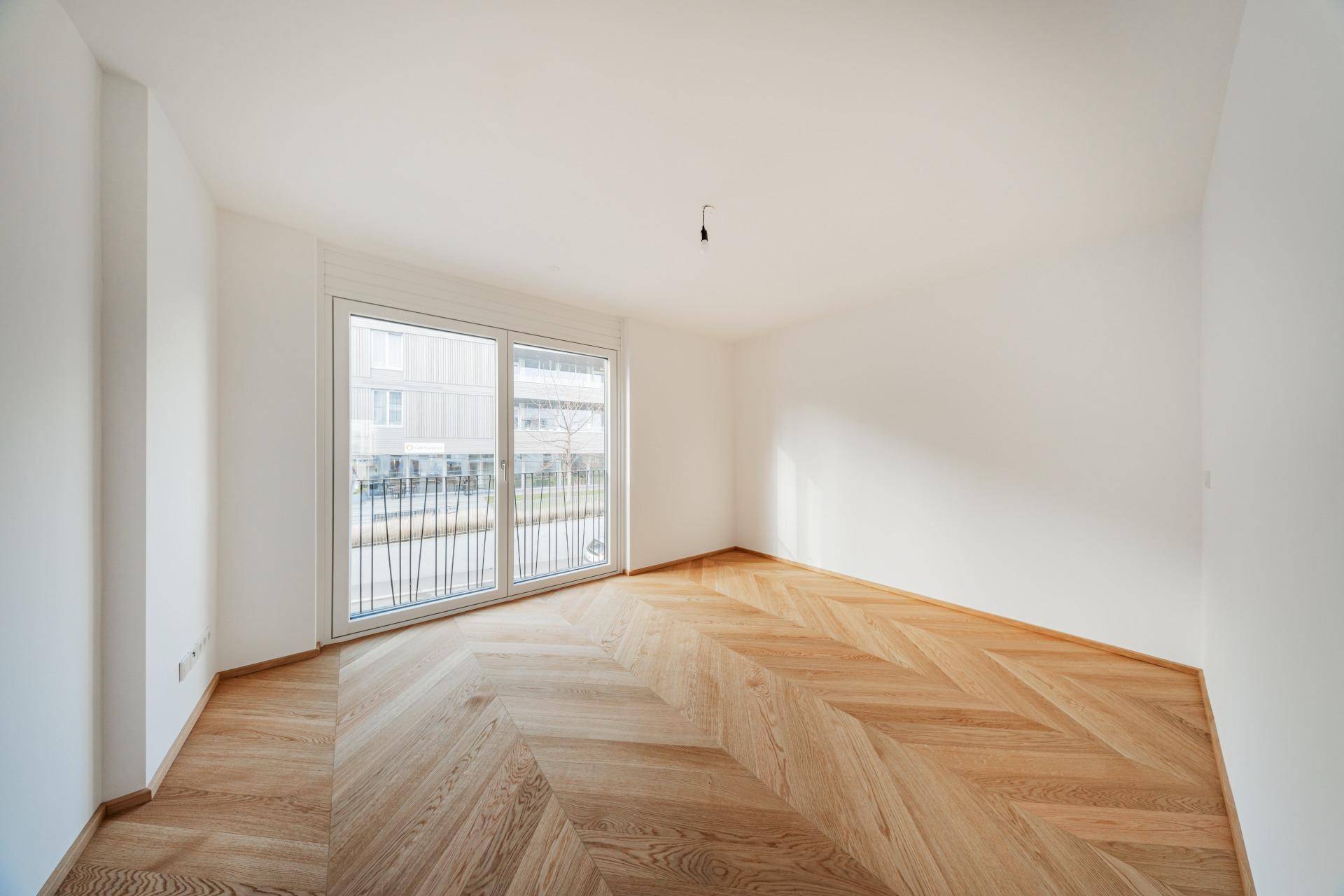 Helles, leeres Schlafzimmer mit modernem Fischgrät-Parkettboden und Zugang zu einem kleinen Balkon.