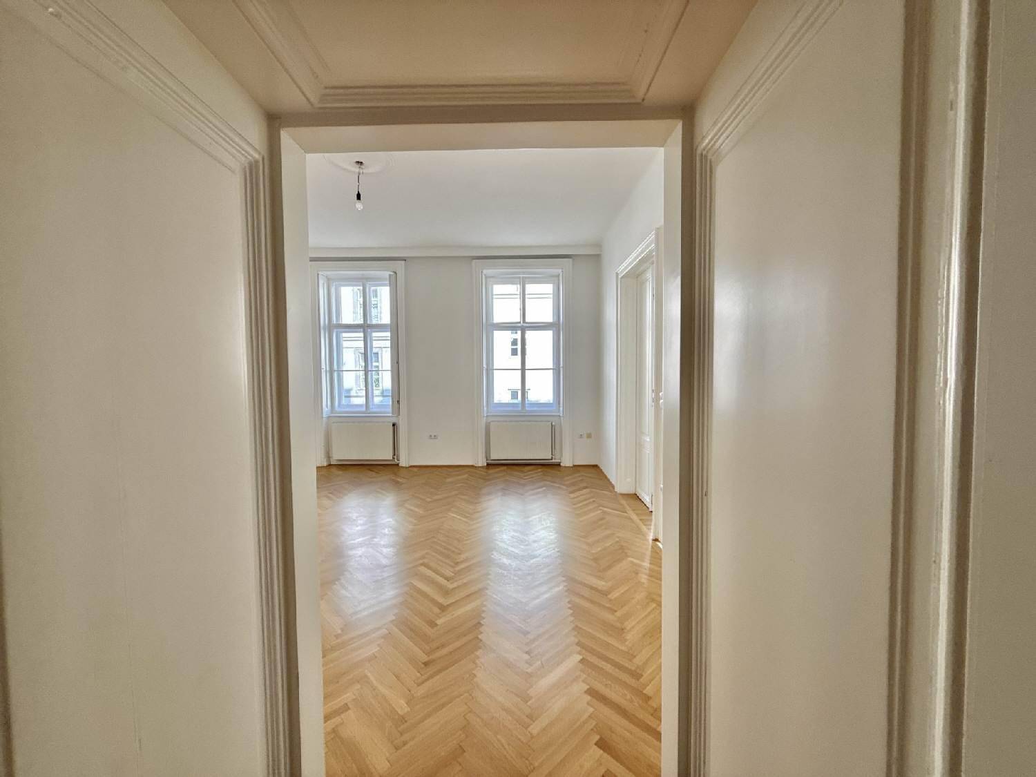 Blick durch eine klassische Doppeltür in ein helles Zimmer mit Fischgrätparkett und hohen Fenstern.