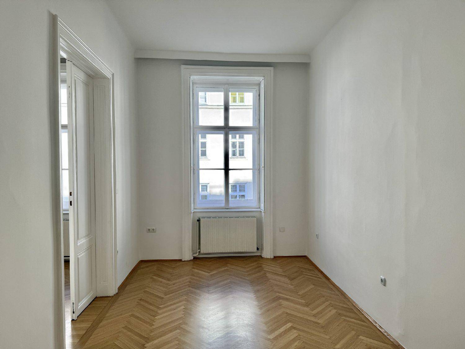 Geräumiges Zimmer mit Fischgrätparkettboden und einem hohen Fenster, das viel Tageslicht in den Raum bringt.