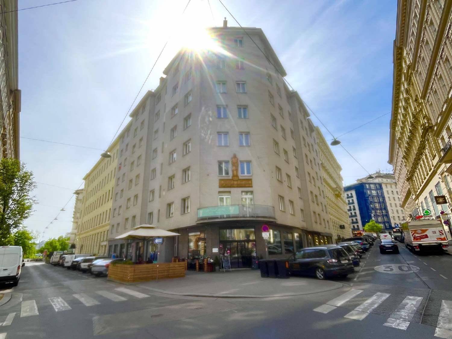 Sonniges Eckgebäude in urbaner Umgebung mit Geschäften im Erdgeschoss und parkenden Autos entlang der Straße.