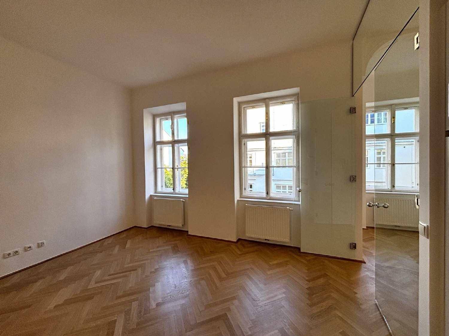 Geräumiger Wohnbereich mit Fischgrätparkett, zwei großen Fenstern und einer eleganten Glaswand.