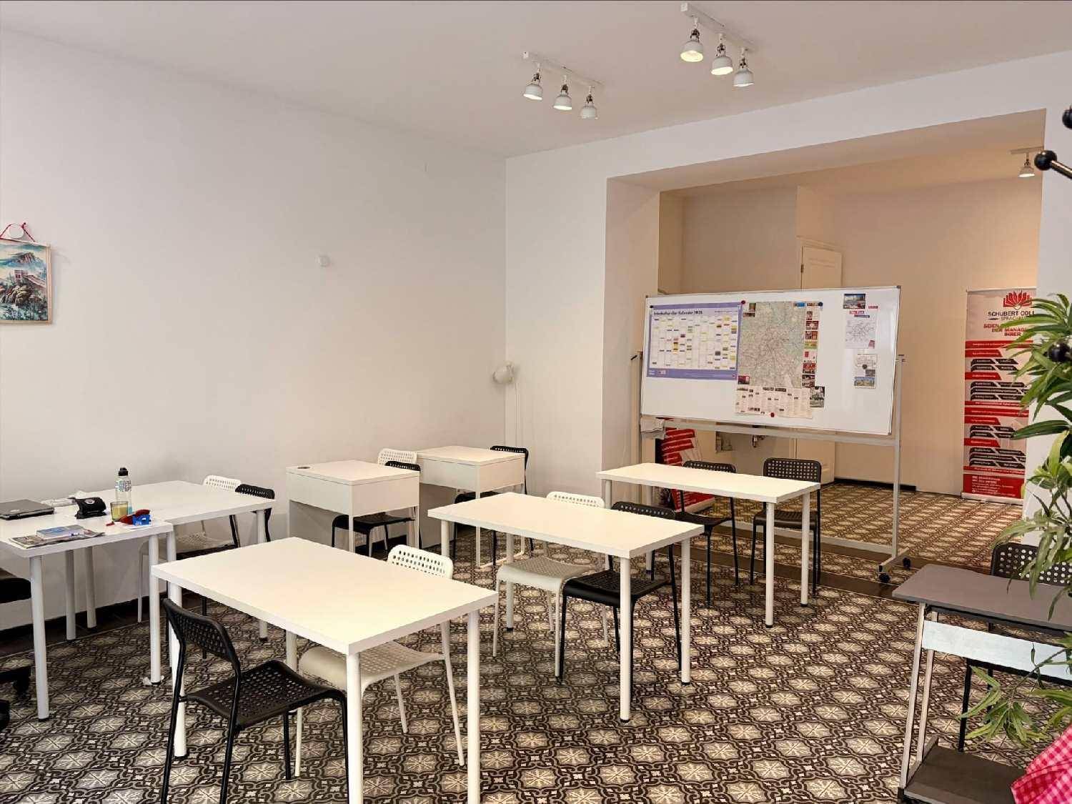 Heller Klassenraum mit mehreren Tischen und Stühlen, einer großen Whiteboard-Tafel und Pflanzen als Dekoration.