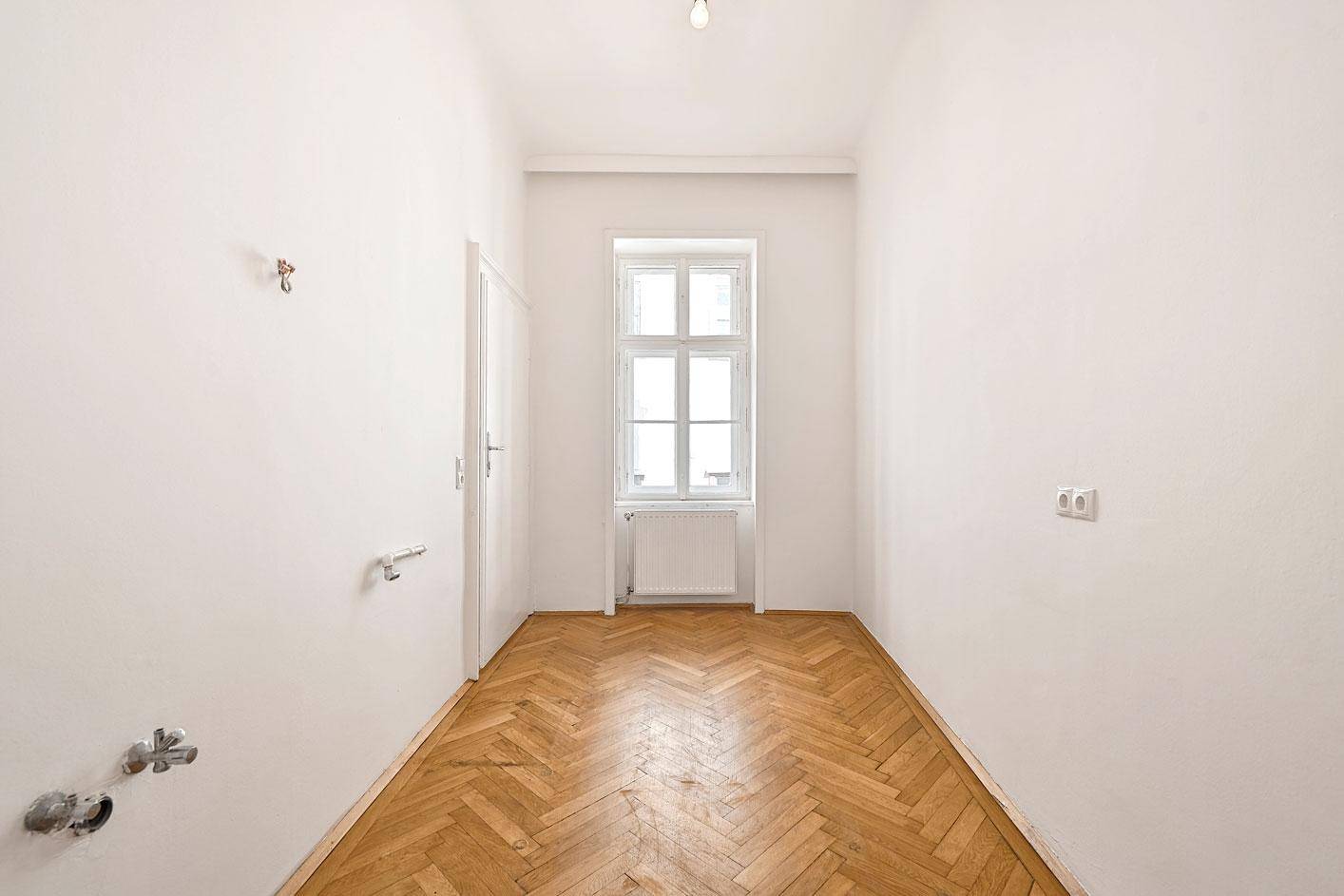 Schmaler Raum mit Fischgrätparkett und einem Fenster, vorbereitet für eine Kücheninstallation.