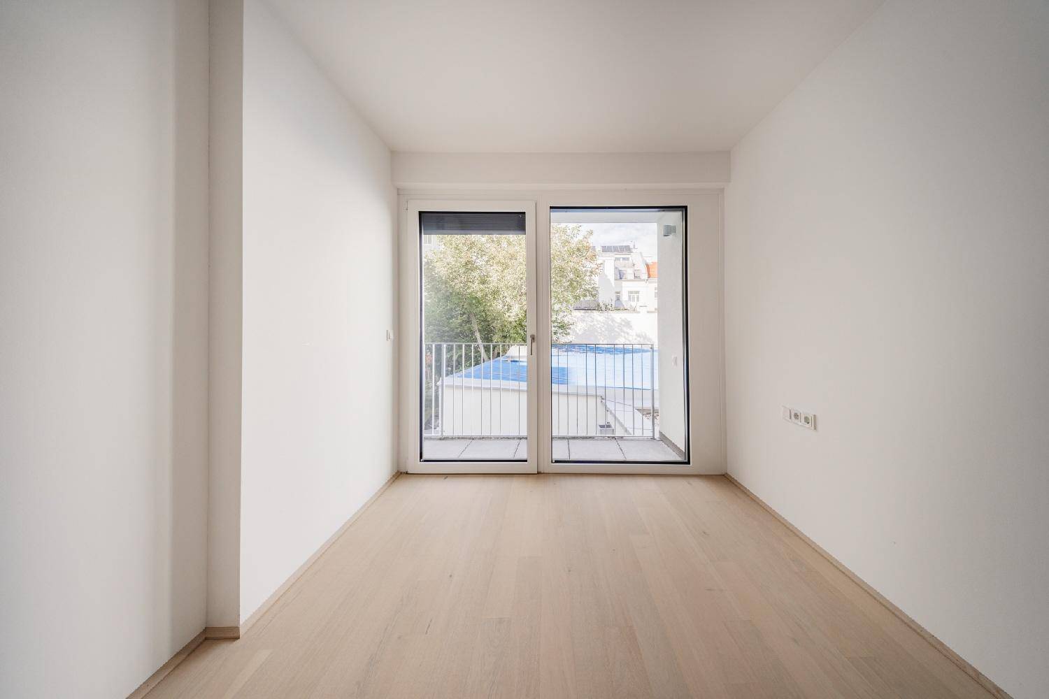 Leeres Zimmer mit Holzboden und Balkonzugang, der einen Blick auf einen Poolbereich bietet.