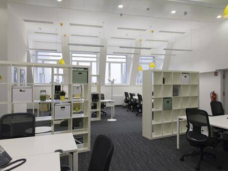 Geräumiges Großraumbüro mit vielen Arbeitsplätzen, Regalen als Raumteiler und großen Fenstern.