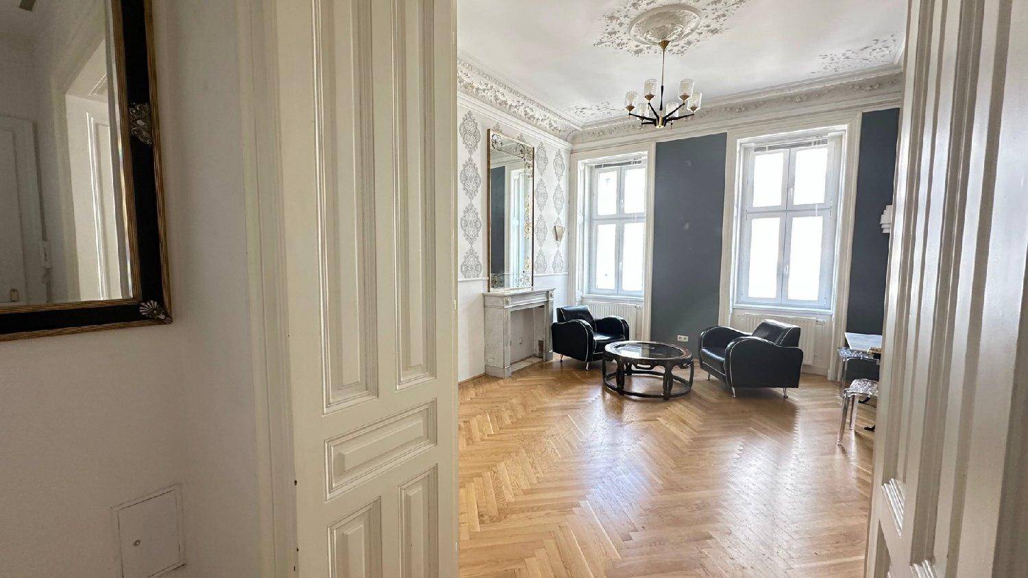 Geräumiges Wohnzimmer mit Fischgrätparkett, Stuckdecke und Blick auf den Flur.