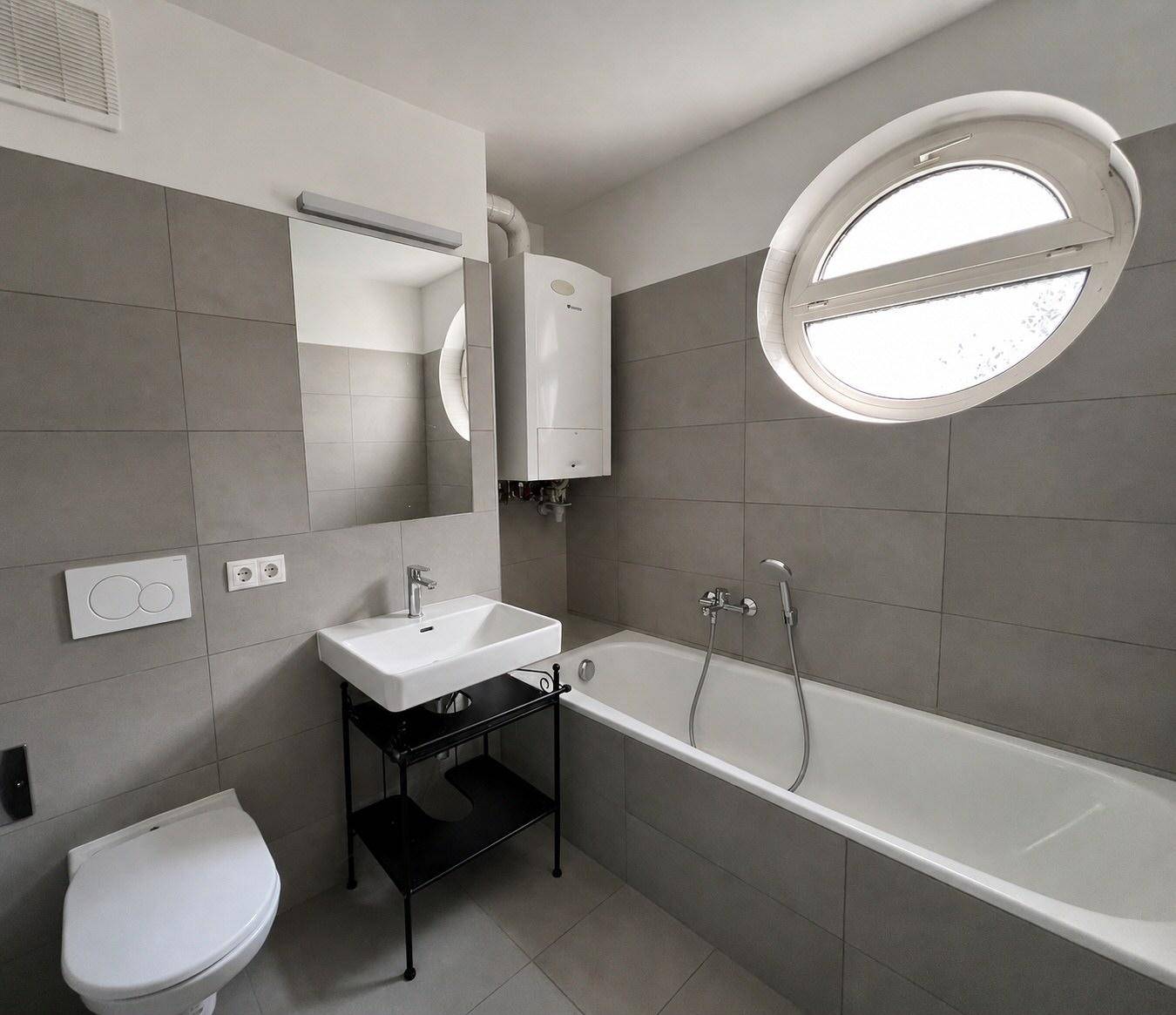 Modernes Badezimmer mit grauen Fliesen, Badewanne, WC und einem ovalen Fenster für Tageslicht.