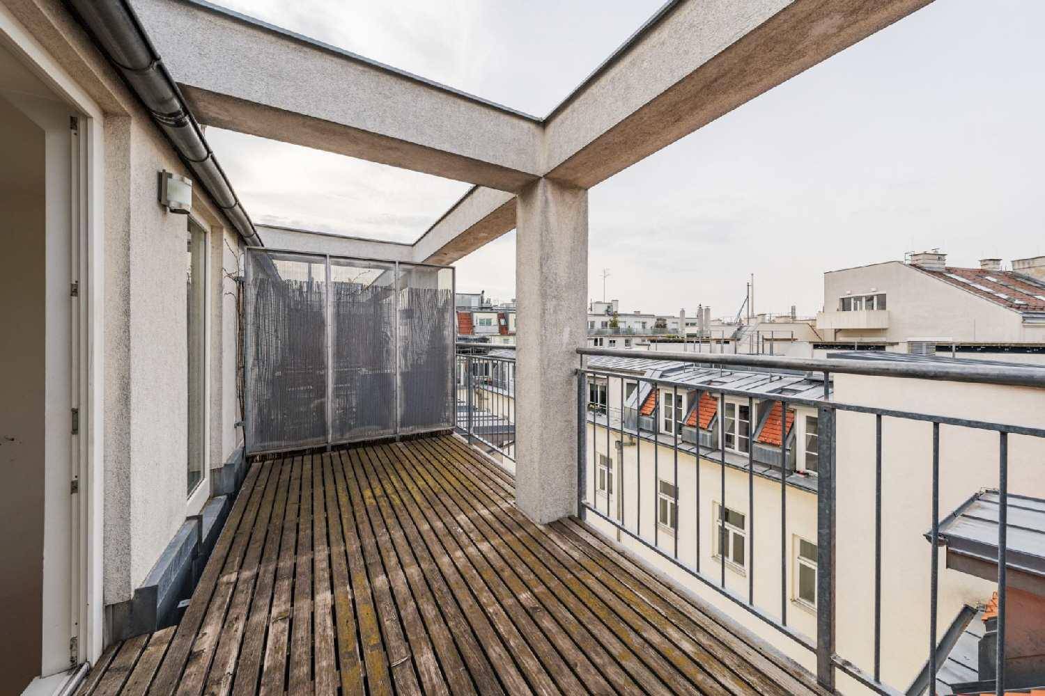 Geräumiger Balkon mit Holzbelag und Blick über die Dächer der Stadt, umgeben von einer Metallbrüstung.