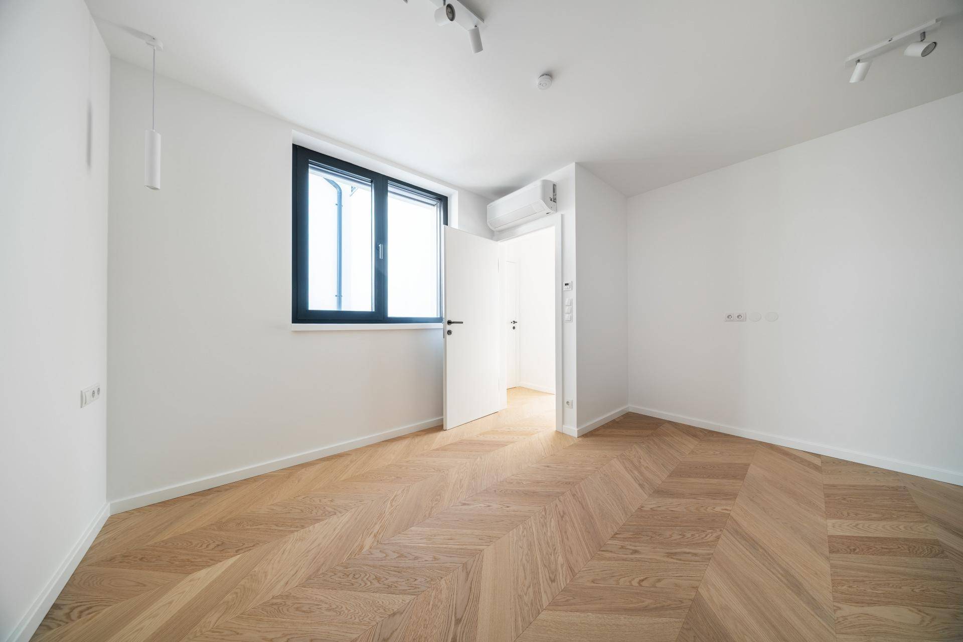 Helles Schlafzimmer mit Fischgrätparkett, Klimaanlage und Fenster, das viel Tageslicht hereinlässt.