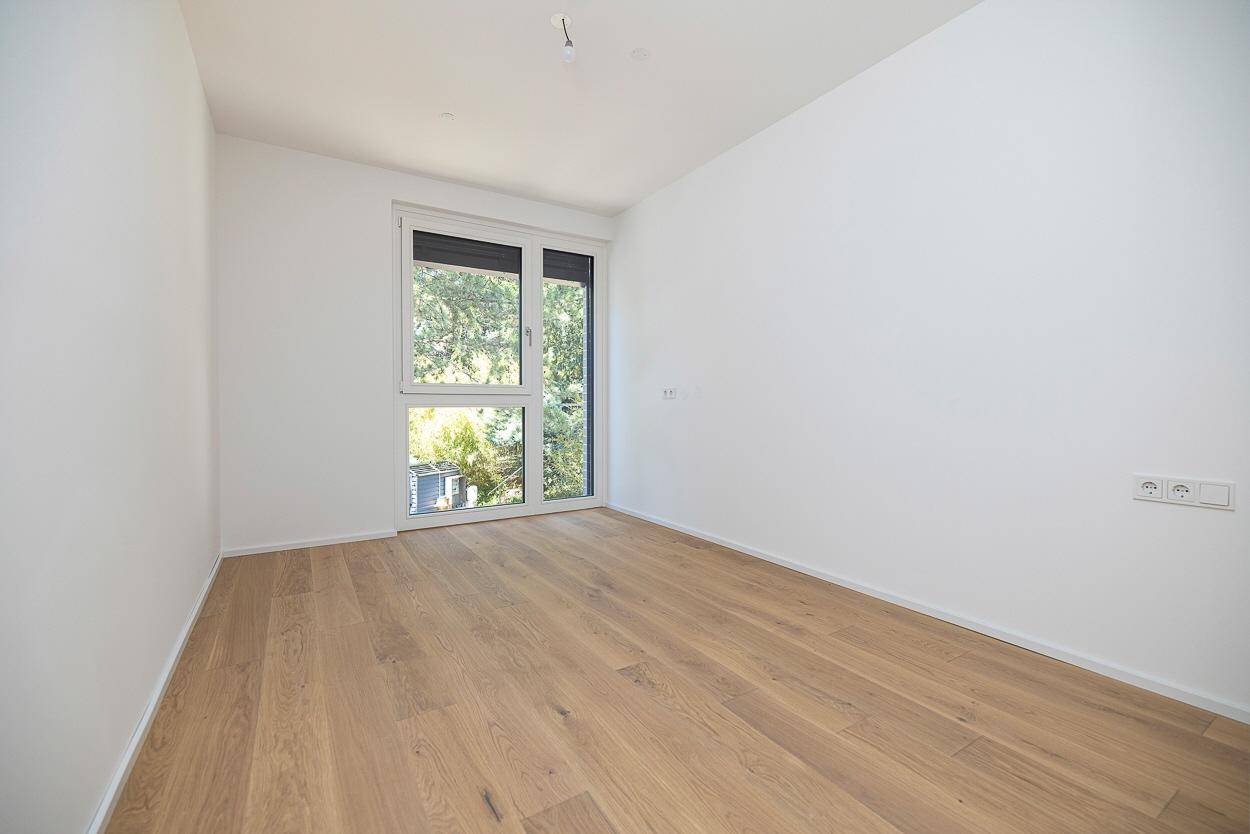 Helles, leeres Zimmer mit Holzboden und großem Fenster mit Blick ins Grüne.