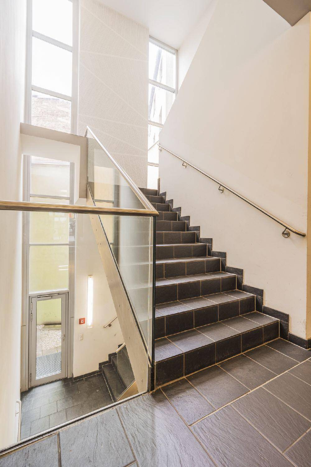 Modernes Treppenhaus mit dunklen Fliesen, Glasgeländer und großen Fenstern für viel Licht.