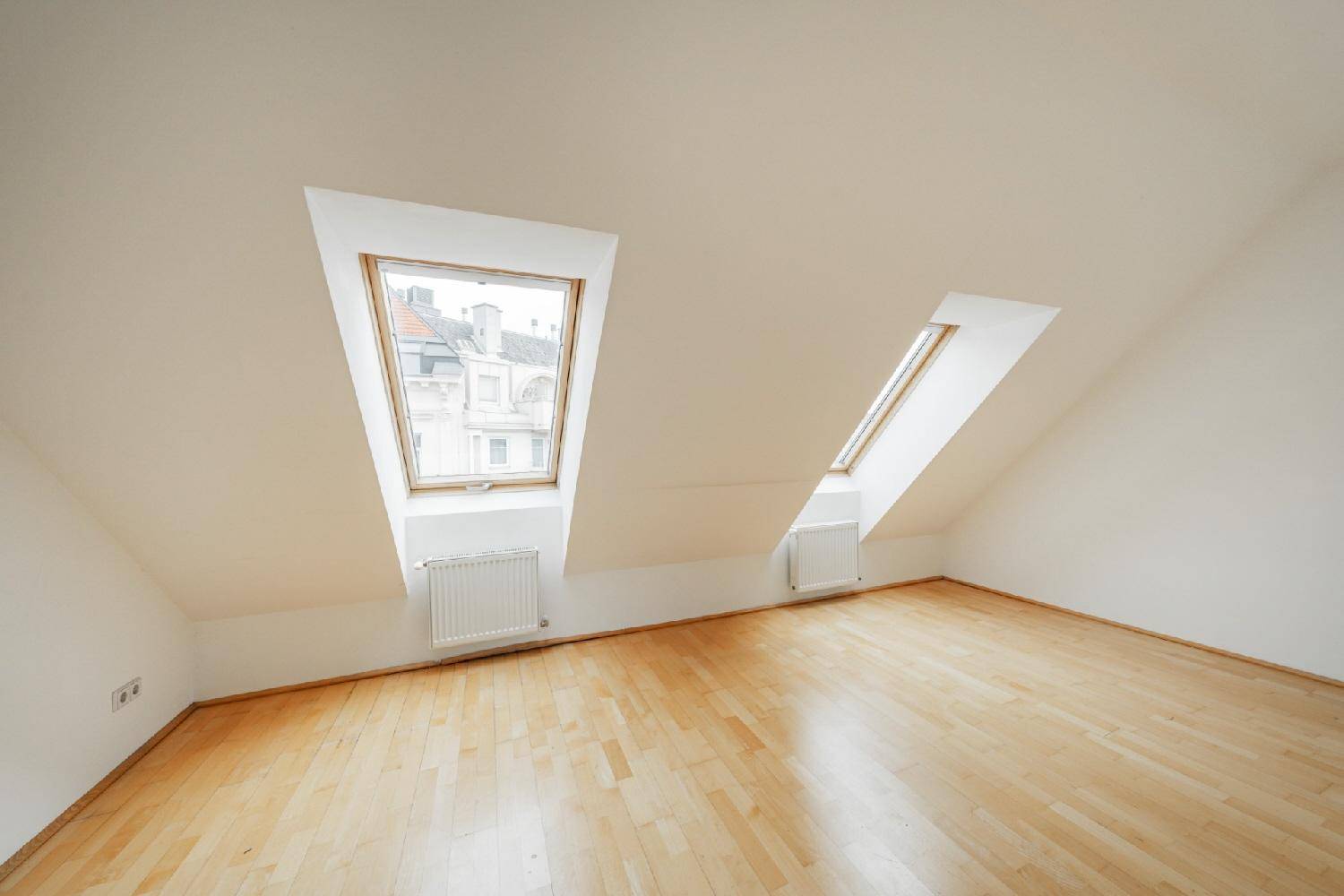Helles Zimmer mit Dachschrägen, Holzfußboden und zwei Dachfenstern, die viel Licht hereinlassen.