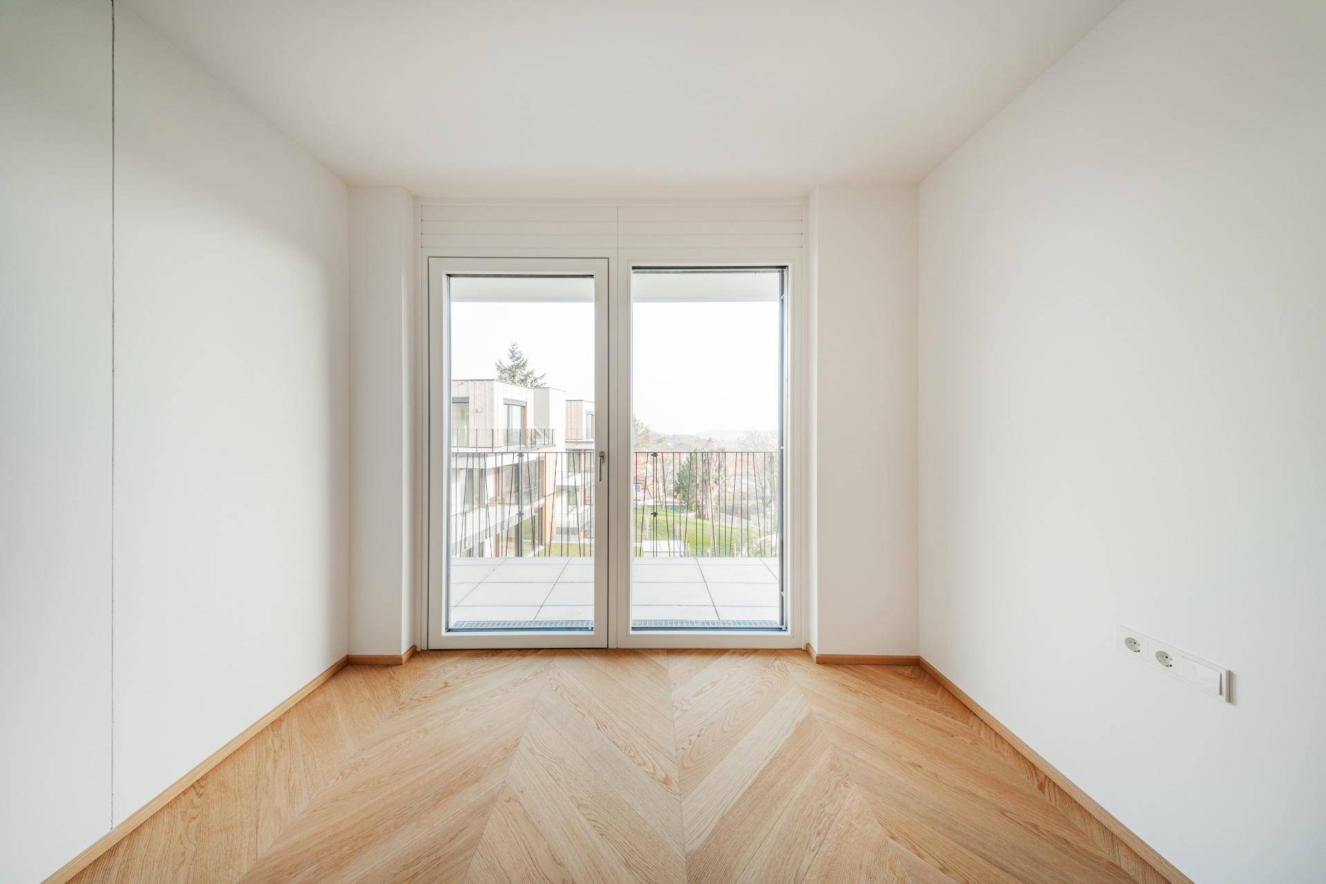 Helles Schlafzimmer mit Fischgrät-Parkettboden und Zugang zu einem kleinen Balkon mit Blick ins Grüne.