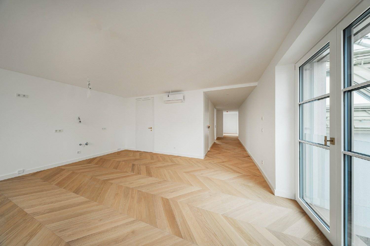 Heller Raum mit Fischgrätparkett, Zugang zu einem Balkon und langem Flur im Hintergrund.