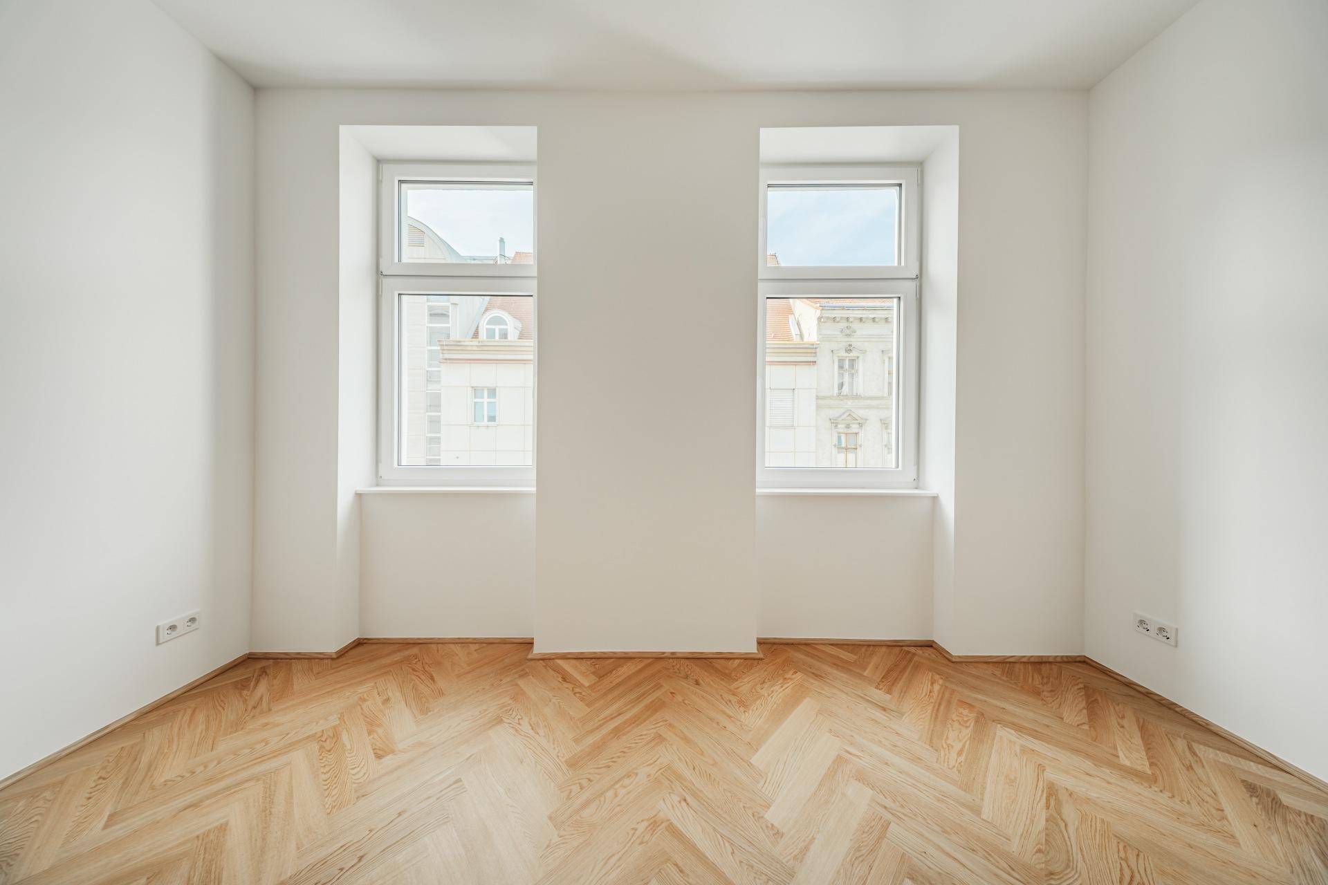 Geräumiges Zimmer mit zwei großen Fenstern, hellem Fischgrätparkett und weißen Wänden.