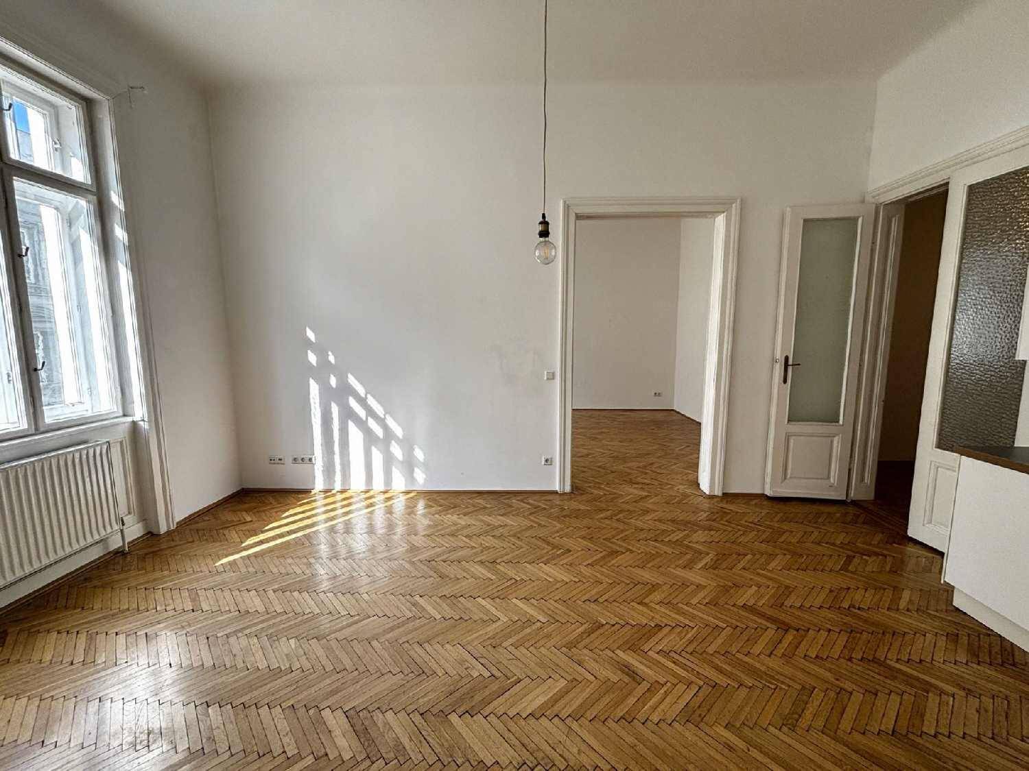 Heller Wohnbereich mit Fischgrätparkett, der zu einem weiteren Zimmer und einer kleinen Küche führt.