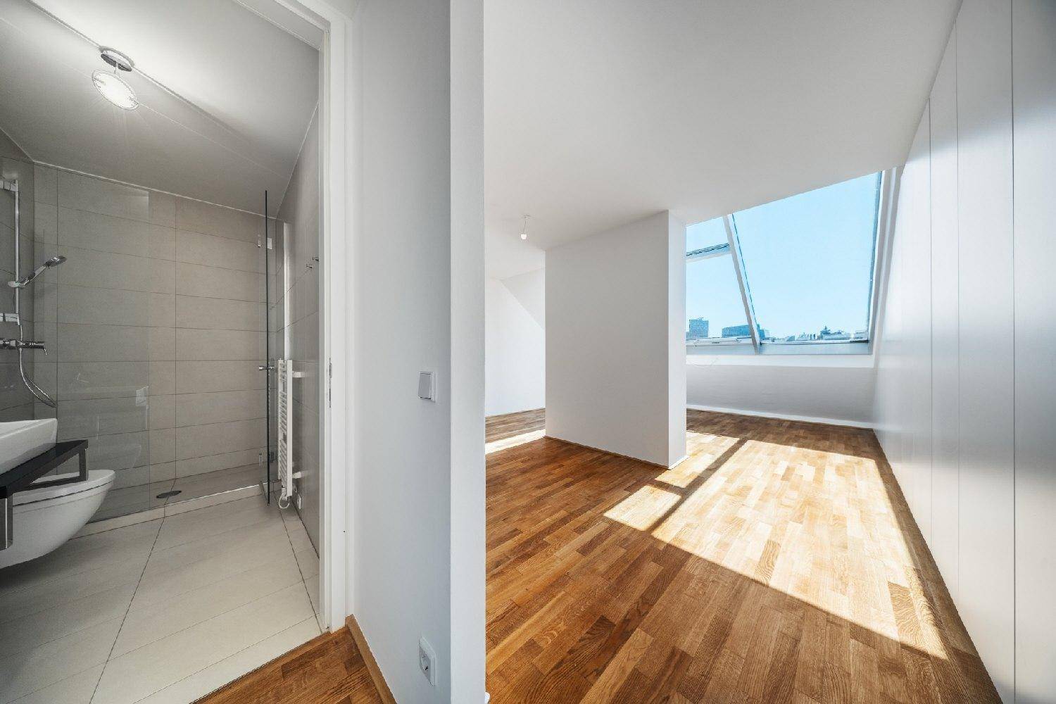 Blick von einem hellen Raum mit Holzboden in ein modernes Badezimmer mit Dusche und WC.