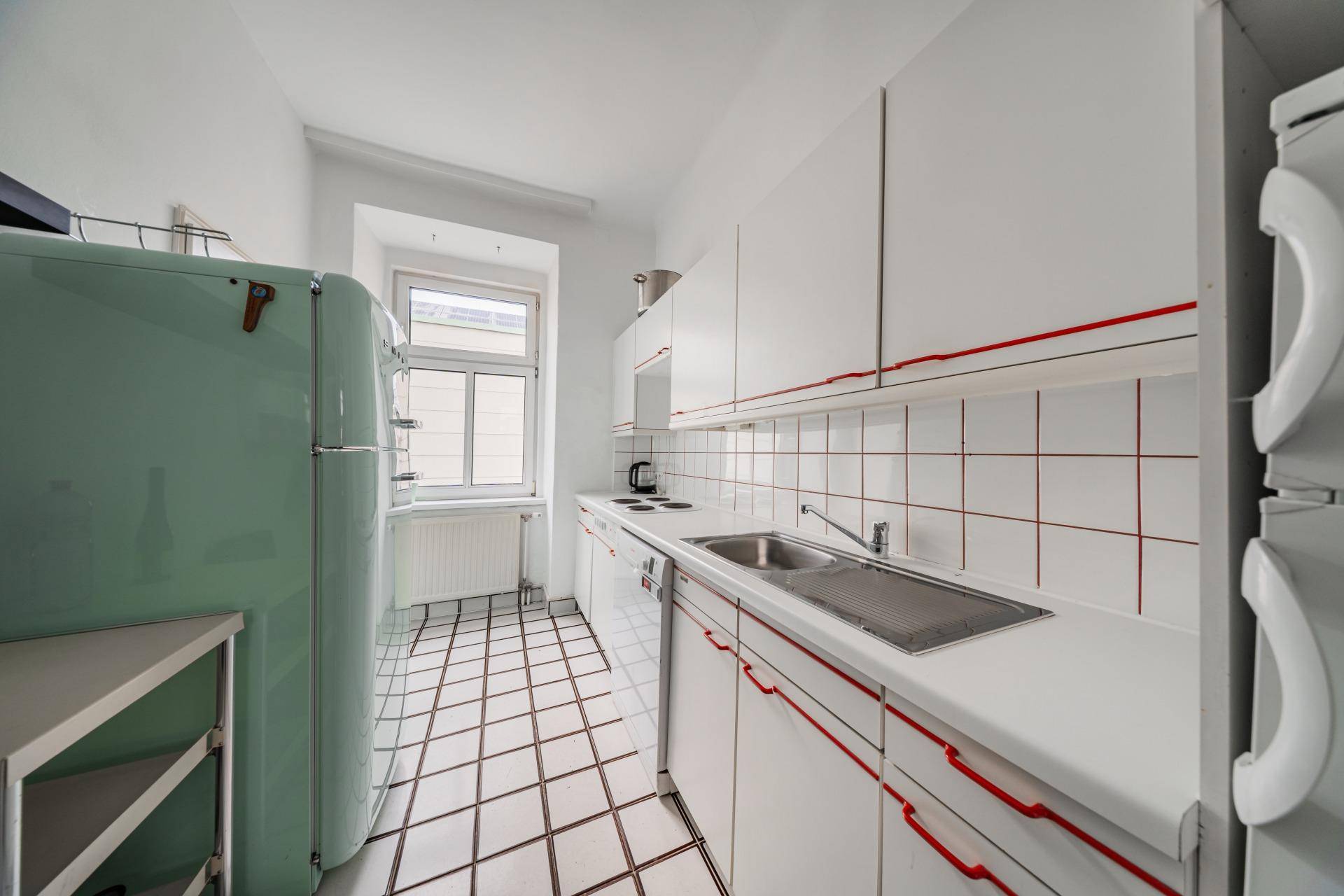 Schmale Küche mit weißen Fliesen, einem mintgrünen Kühlschrank und einem Fenster.
