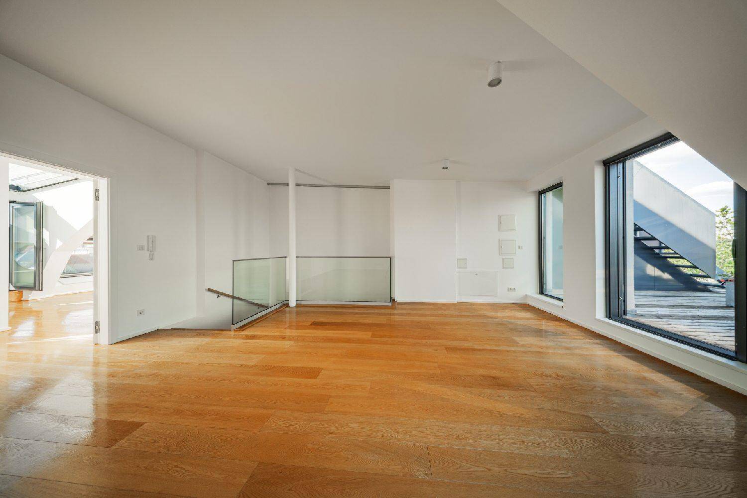 Heller Wohnraum mit Holzboden, Glasgeländer und großen Fenstern, die den Blick nach draußen freigeben.
