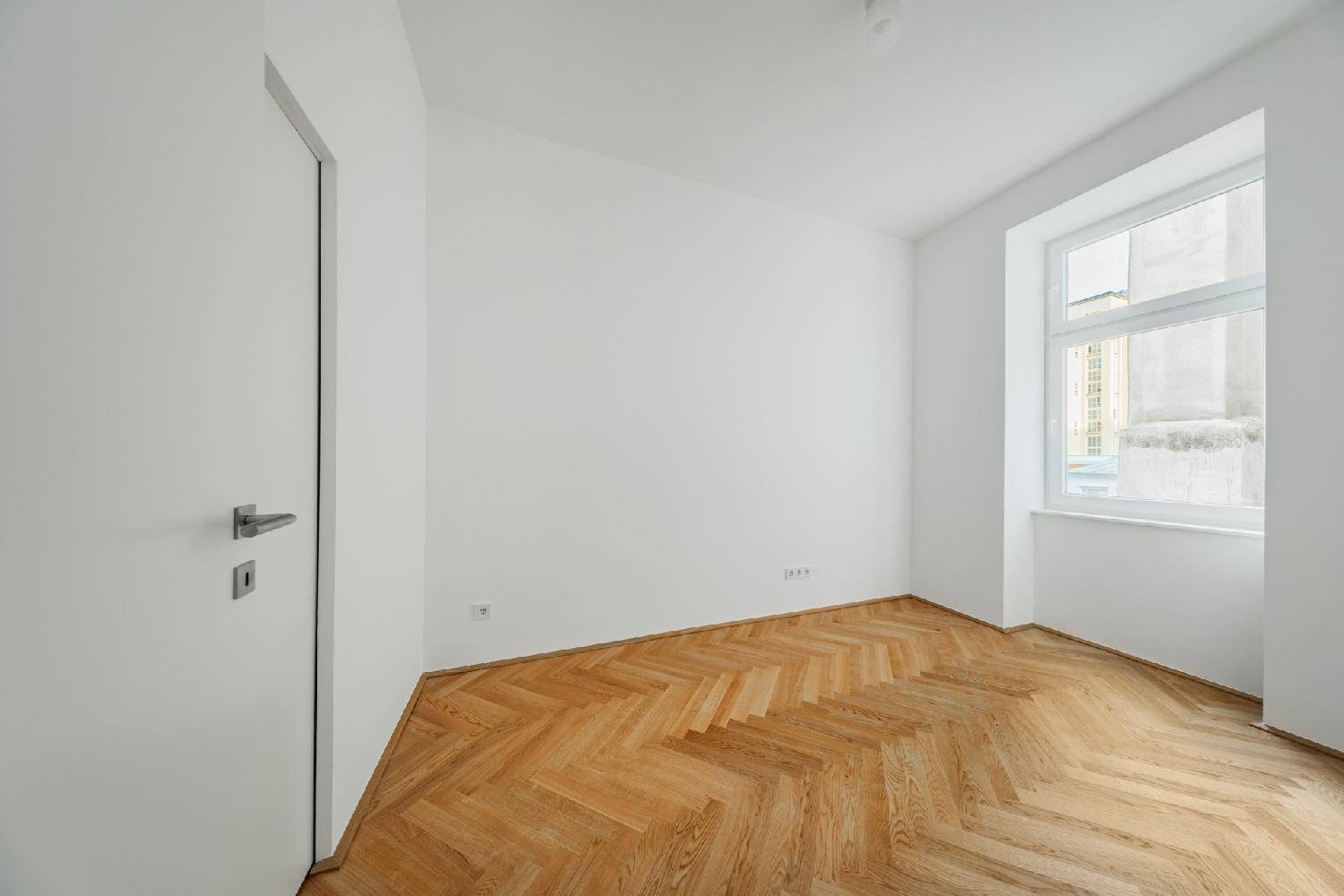 Leeres Zimmer mit Fischgrätparkett und einem Fenster, ideal als Schlaf- oder Arbeitszimmer.