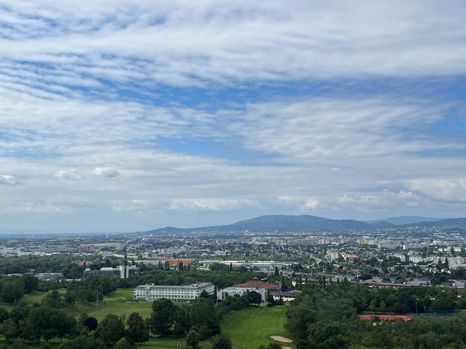 Weitläufiger Panoramablick über eine Stadtlandschaft mit viel Grün und Bergen am Horizont unter blauem Himmel.