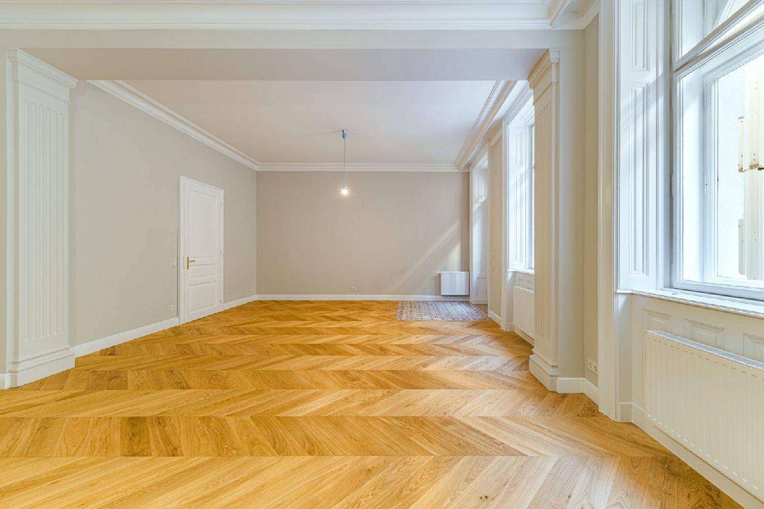 Großer, heller Wohnraum mit Fischgrätparkett, hohen Decken und klassischen Fenstern, die viel Licht spenden.
