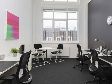 Helles Großraumbüro mit mehreren Arbeitsplätzen, großen Fenstern und dunklem Teppichboden für Komfort.
